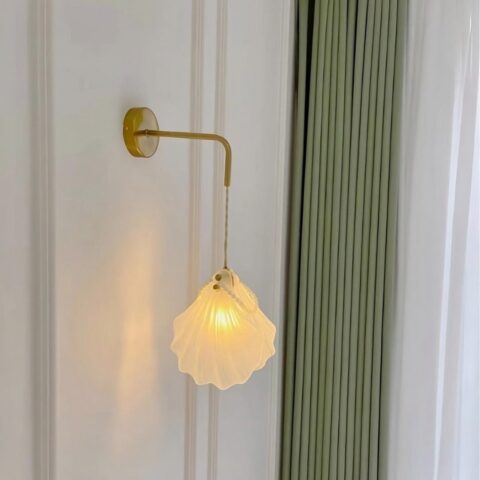 Applique murale coquillage en verre et perles accrochée sur un mur blanc avec un rideau vert à côté.