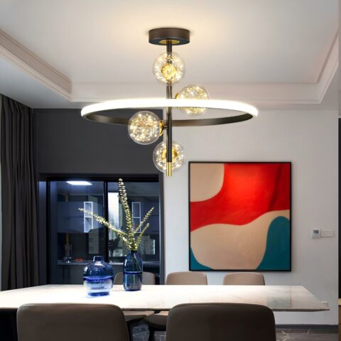 Lustre avec support rond noir au plafond, une verticale noire fixée dessus. 4 boules transparentes avec fils dorées à l'intérieur et diodes de lumière donnant l'impression que ça scintille. L'ensemble est entouré d'un cercle lumineux à l'extérieur et noir à l'intérieur. Fixé au-dessus d'une table rectangulaire blanche avec 5 chaises grises autour. 2 vases bleus sur la table remplis d'eau dont un à des fleurs longues. Sur le mur en face, un cadre abstrait beige, rouge et bleu. A gauche une baie vitrée avec mur gris, encadrement noir et rideau gris.