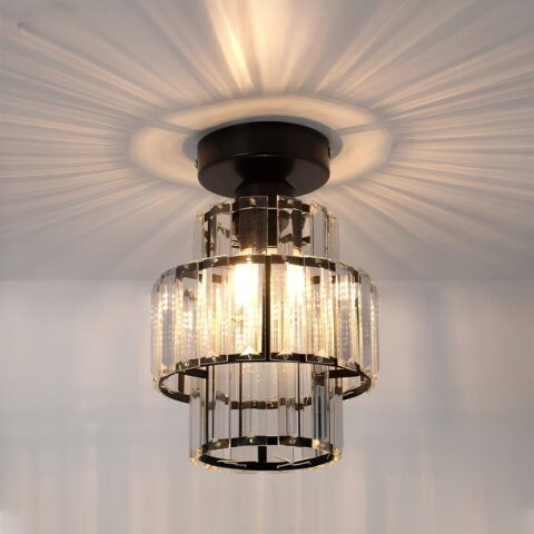 Lustre plafonnier de forme cylindrique avec 3 rangées de lamelles style cristal. Le support u plafond est de couleur noire et les lamelles sont fixées sur des cercles noirs. Les ampoules sont allumées et diffusent des rayons au plafond autour du lustre.