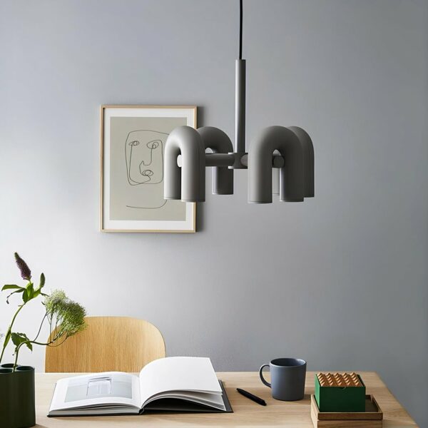 Lustre gris avec 4 éléments en forme de U à l'envers au-dessus d'un bureau beige avec un livre ouvert, une tasse , une plante à gauche, une chaise beige. Derrière un mur gris clair avec un cadre.
