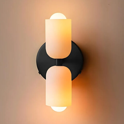 Applique murale à double éclairage, un cylindre en verre vers le haut et un vers le bas fixés sur un socle rond noir. Les ampoules sont allumées.