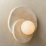 Applique murale circulaire en pierre naturelle de couleur beige avec deux ronds en pierre l'un sur l'autre. Au centre, une boule lumineuse. L'applique est fixée sur un mur beige.