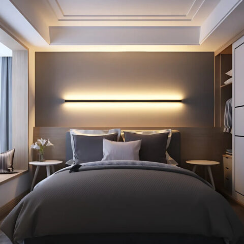 Applique murale minimaliste noire en forme de tube horizontal allumé avec la lumière chaude diffusée au-dessus et en-dessous. Elle est fixée dans une chambre au-dessus d'un lit deux places avec une literie grise.