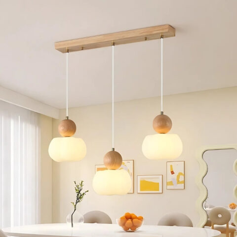 Lustre 3 suspensions rondes alignées en bois clair et acrylique. Les 3 suspensions sont suspendues sur une barre en bois fixée au plafond. Le lustre se trouve au-dessus d'une table blanche avec des sièges gris. Une fenêtre sur la gauche et un mur beige à l'arrière.