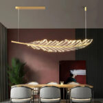 Lustre en forme de feuille suspendue en acrylique avec un support fixé au plafond de couleur doré. Il est au-dessus d'une table rectangulaire en bois avec 6 chaises grises. Au fond un mur noir et bordeaux avec un tableau peint. Sur la gauche, une plante style palmier et un rideau gris.