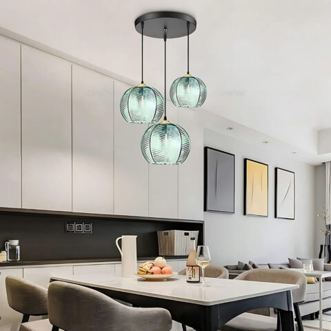 Suspension 3 lampes en verre de couleur verte avec un motif de feuilles. Chaque globe est suspendu par un câble noir. La suspension est au-dessus d'une table rectangulaire. Sur la droite, un canapé gris avec trois cadres colorés au-dessus.