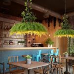 Suspension jungle avec feuillage suspendue au-dessus d'une table dans un établissement tel qu'un restaurant. La suspension est allumée et de forme ronde. La table en-dessous est carrée et en bois avec deux chaises autour. En arrière plan, un conmptoir de bar.
