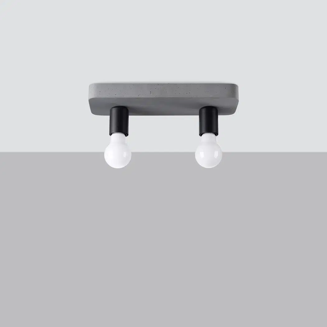 Plafonnier rectangulaire en béton gris fixé au plafond. Deux ampoules blanches de forme classique pendent vers le bas, chacune insérée dans une douille cylindrique noire. L’ensemble a un style très simple et minimaliste, avec un côté brut et industriel. L’arrière-plan est neutre, gris clair, ce qui met en valeur la sobriété du luminaire.