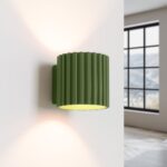 Applique murale vert olive de forme cylindrique et texturée, sur un mur blanc avec une fenêtre en fond