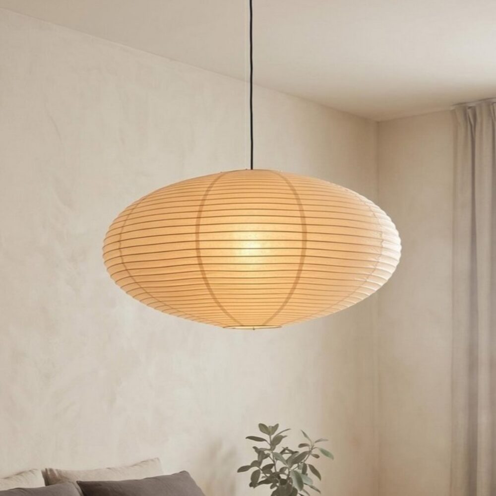 Une Suspension Japandi ellipse en papier de riz en forme d’ovale, lumineuse et douce, suspendue dans une pièce au mur beige avec un canapé et une plante en dessous.