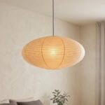 Une Suspension Japandi ellipse en papier de riz en forme d’ovale, lumineuse et douce, suspendue dans une pièce au mur beige avec un canapé et une plante en dessous.