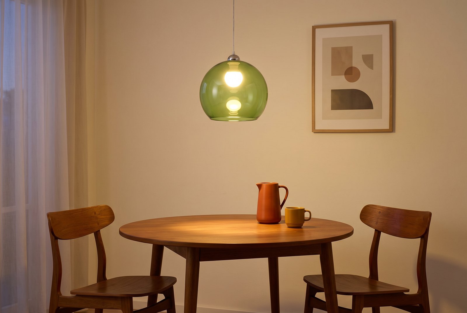 Coin repas mid century le soir, suspension globe en verre vert allumée au-dessus d'une table ronde en teck, deux chaises en bois, pichet orange et tasse moutarde