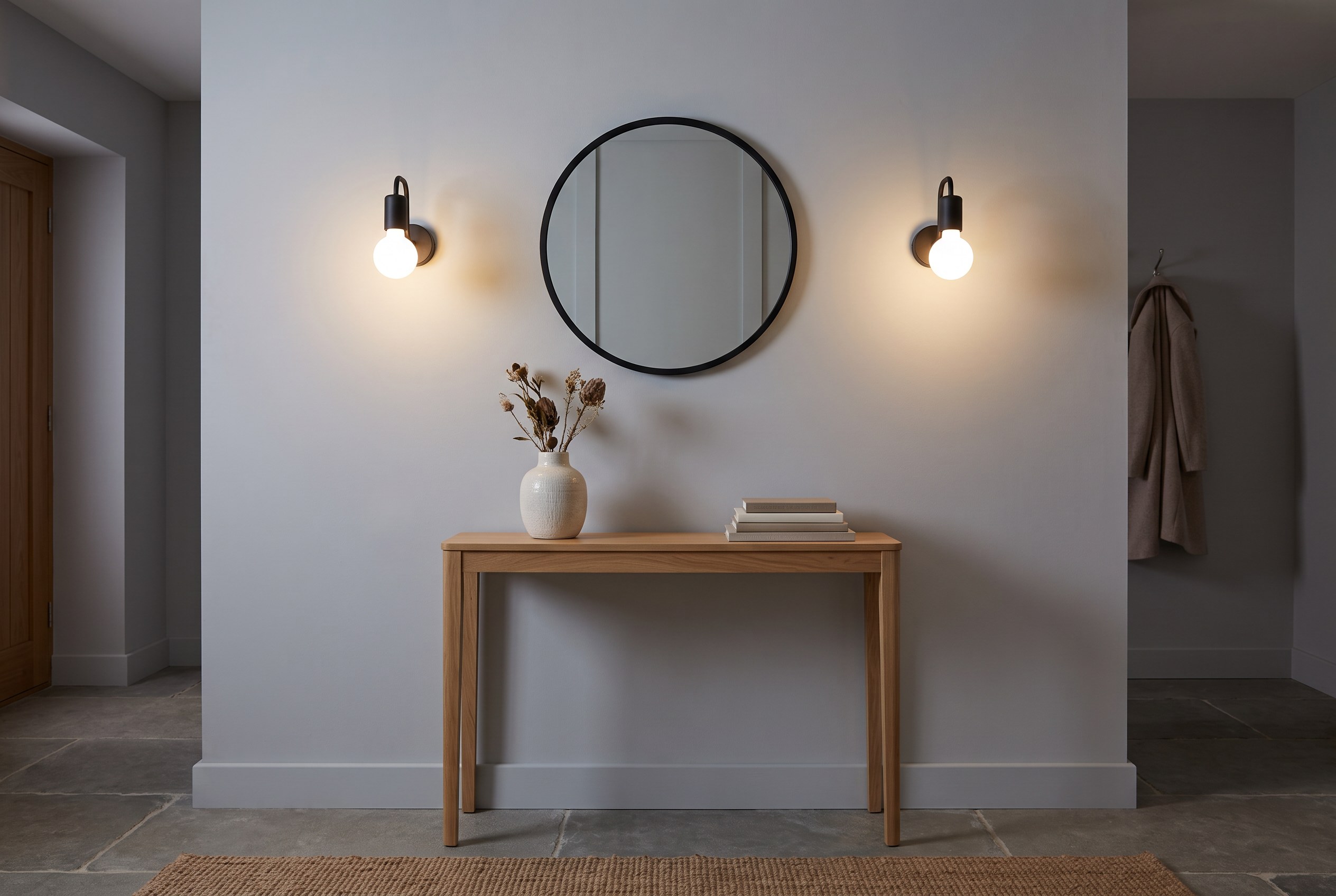 Entrée aux murs gris clair avec deux appliques murales en acier noir à ampoule globe allumées de part et d'autre d'un miroir rond, au-dessus d'une console en chêne