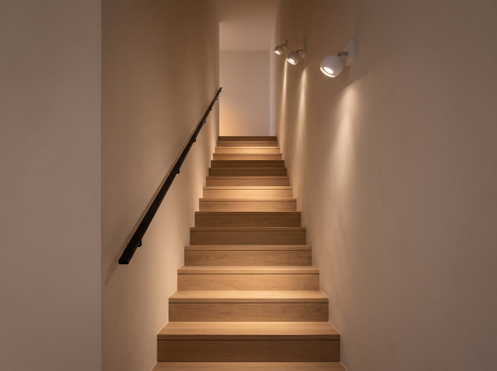Escalier droit étroit en chêne entre deux murs clairs, éclairé par trois appliques à spot rond orientable blanc dont les têtes sont tournées vers les marches