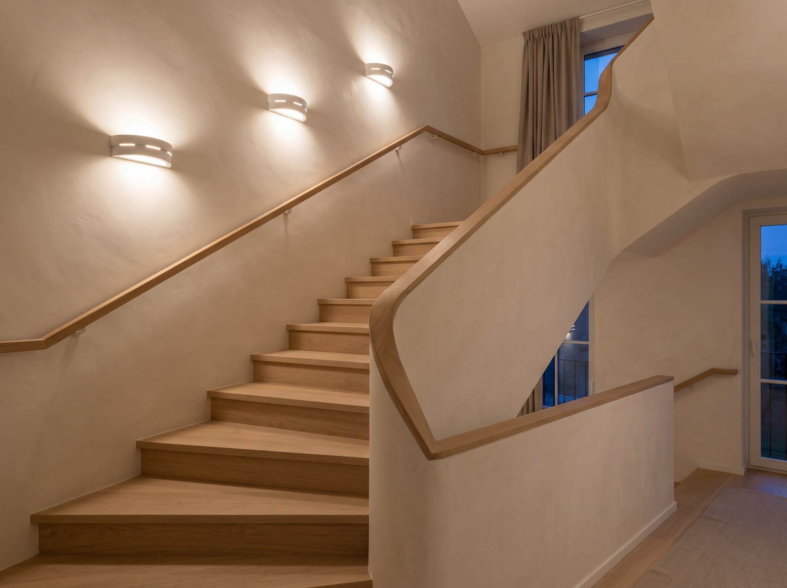 Escalier quart tournant en chêne le soir, avec trois appliques murales demi-lune en céramique blanche allumées le long de la pente, lumière vers le haut et vers le bas sur un mur blanc cassé