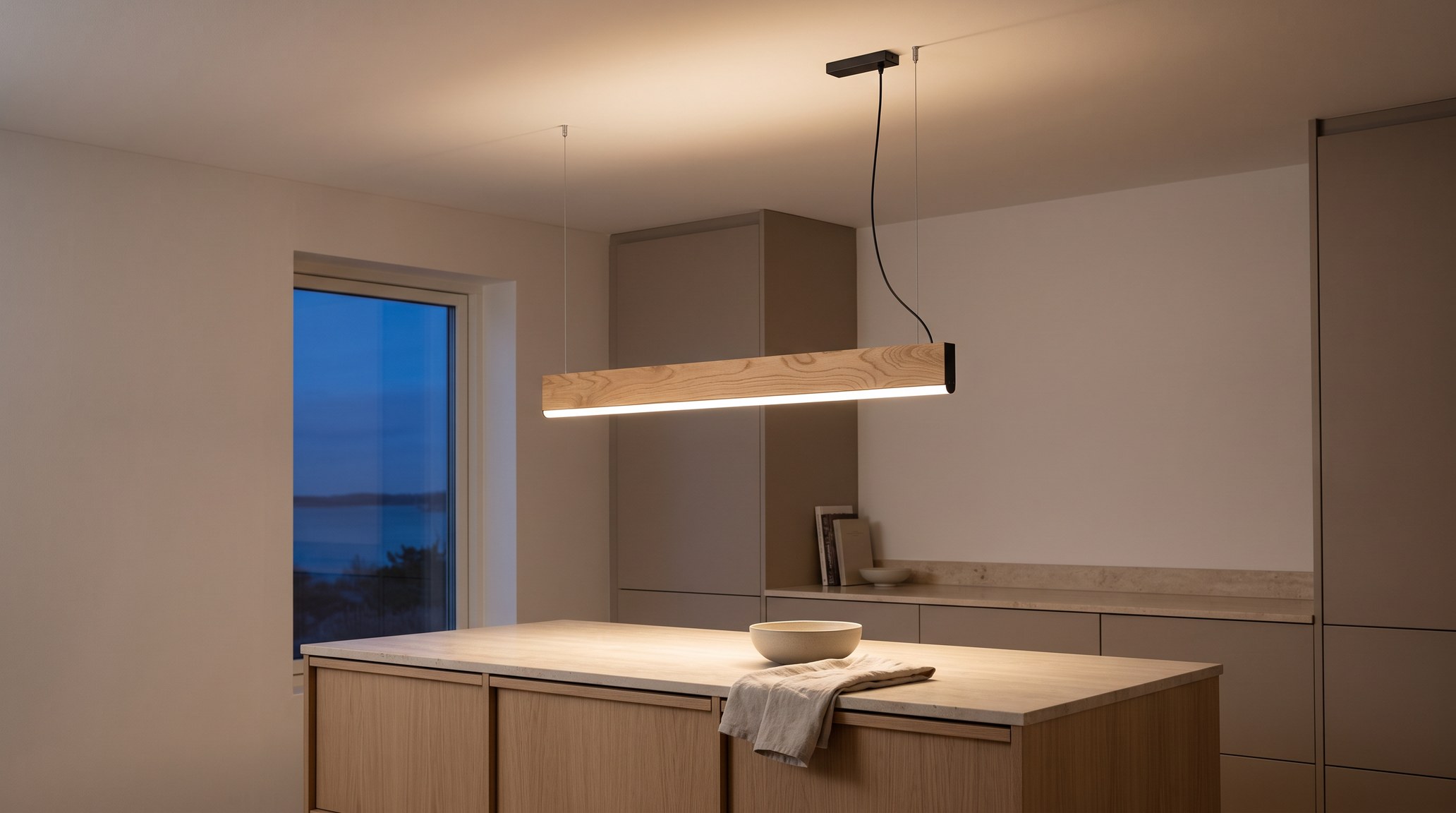 Suspension linéaire LED en chêne clair à diffuseur blanc allumée le soir au-dessus d'un îlot de cuisine en pierre claire, cuisine aux façades taupe