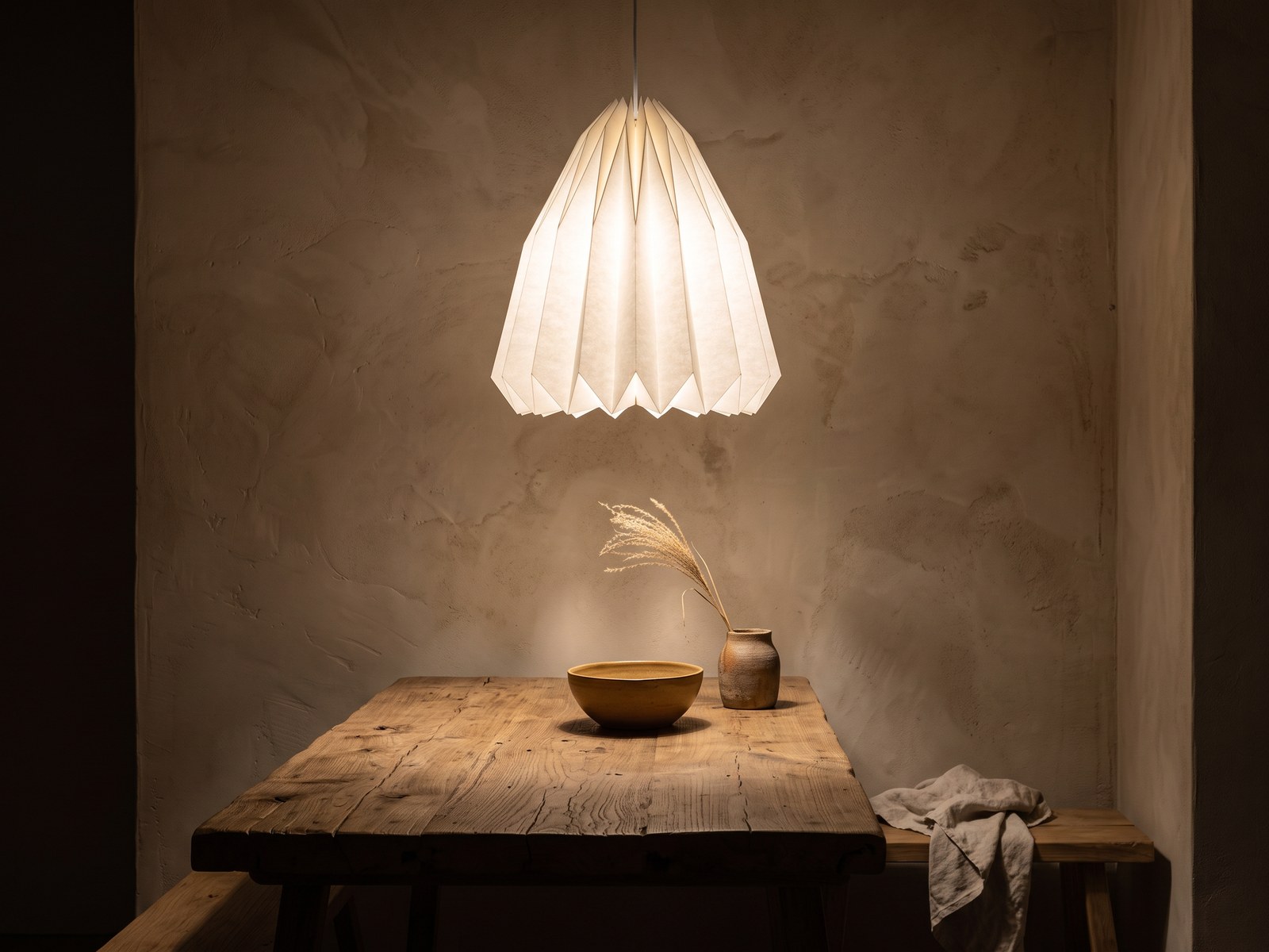 Coin repas wabi-sabi le soir, suspension en papier blanc plissé allumée en blanc chaud au-dessus d'une table en bois brut, mur enduit à la chaux et bol en céramique, reste de la pièce dans la pénombre