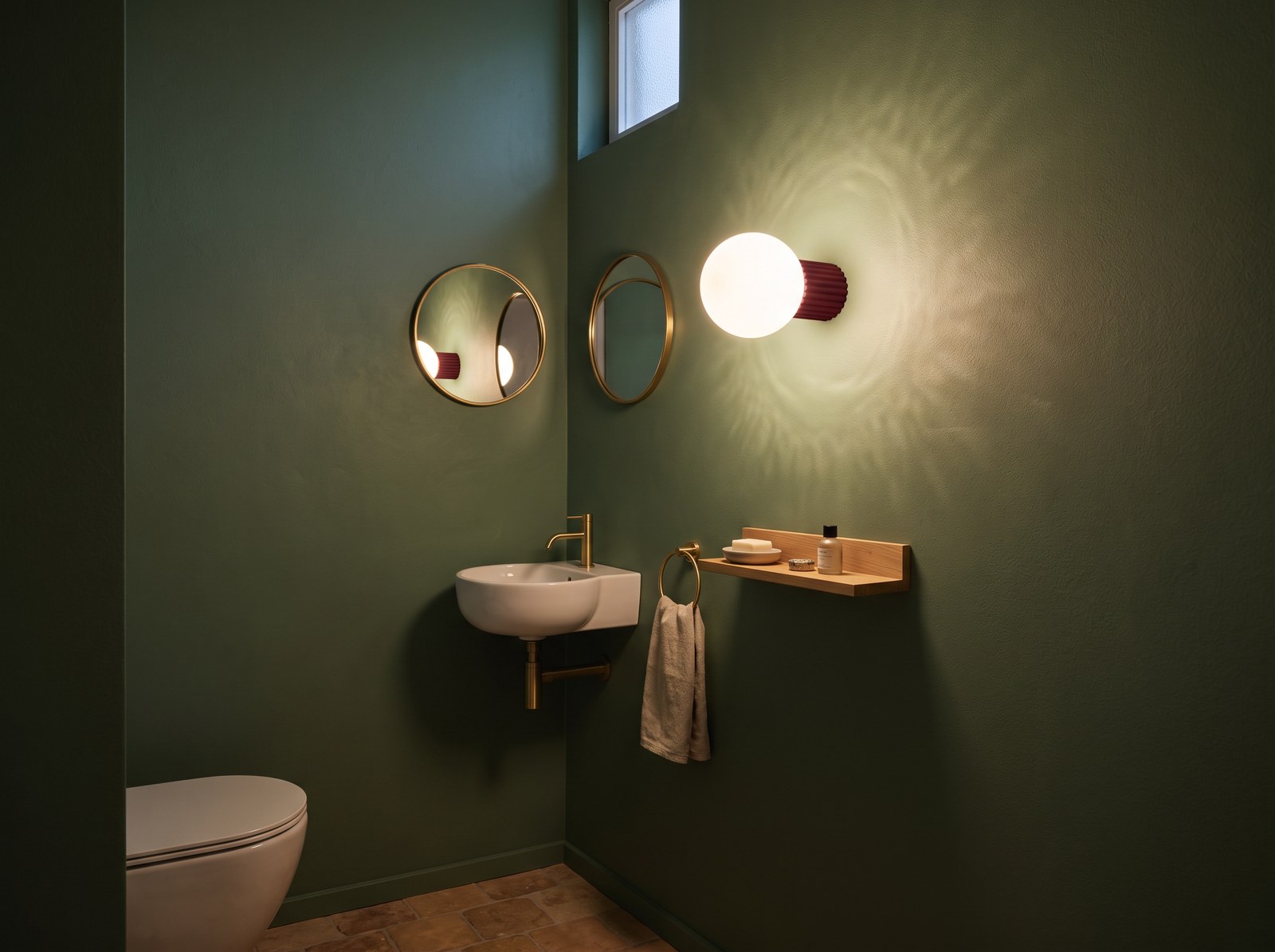 Applique murale à globe opalin et base rainurée rouge bordeaux allumée au-dessus d'un petit lave-mains dans des toilettes aux murs vert sauge